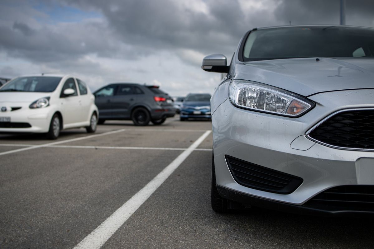 A silver car parked in between the lines of a parking lot. There is a black car driving away from the parking spots.