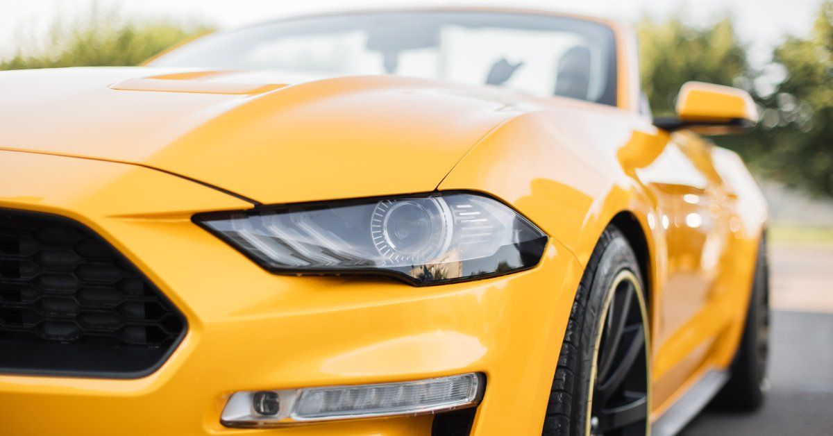 A close-up of a yellow convertible sports car parked outside. Its front bumper and the headlight of the car are in the foreground.
