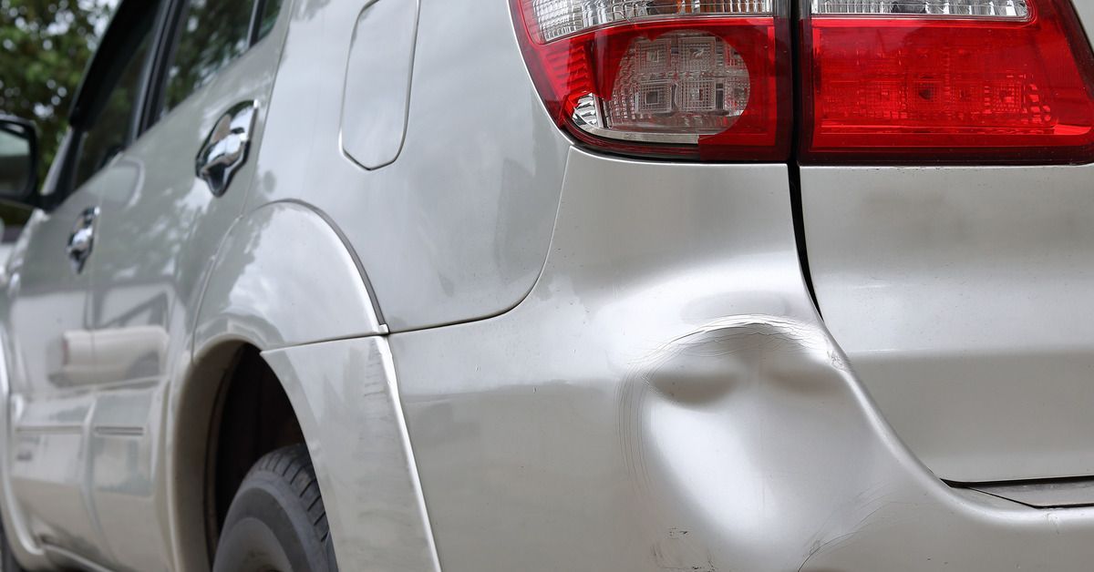 A silver car parked in an outdoor lot. There is a small dent on the bumper on the back left side of the vehicle.