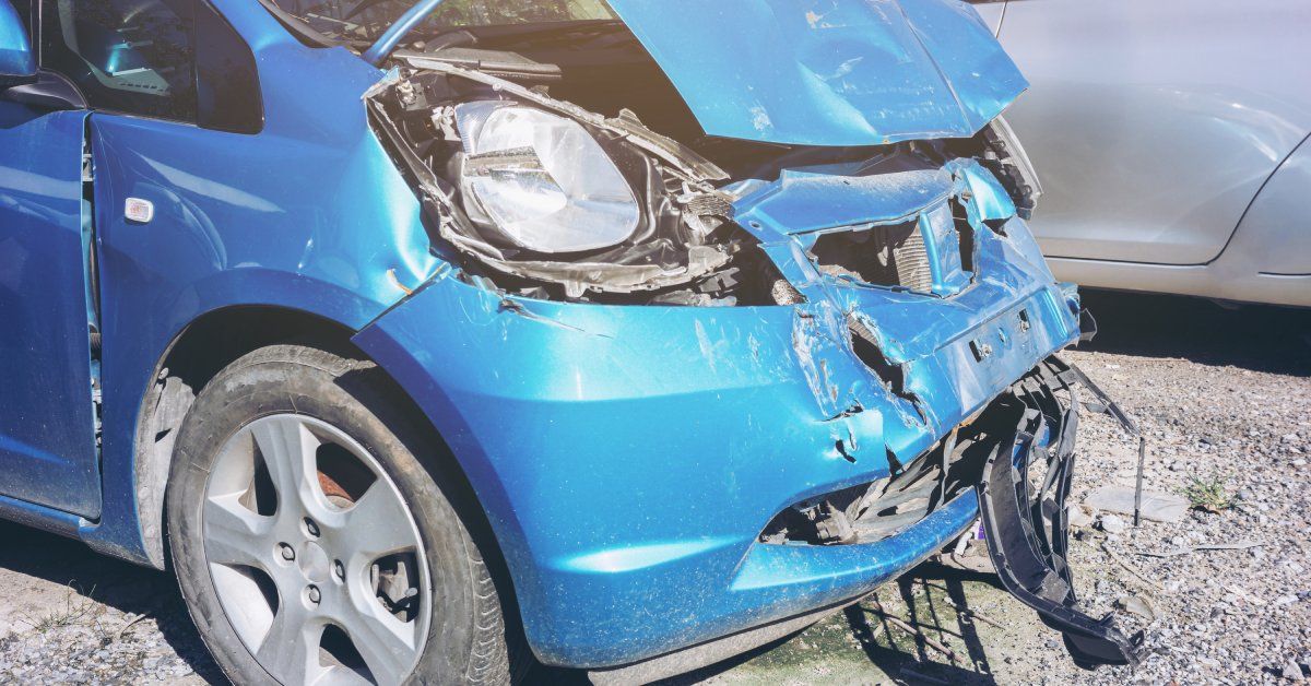 A blue vehicle with its front bumper and hood crumpled due to a head-on collision. There is a silver car nearby.