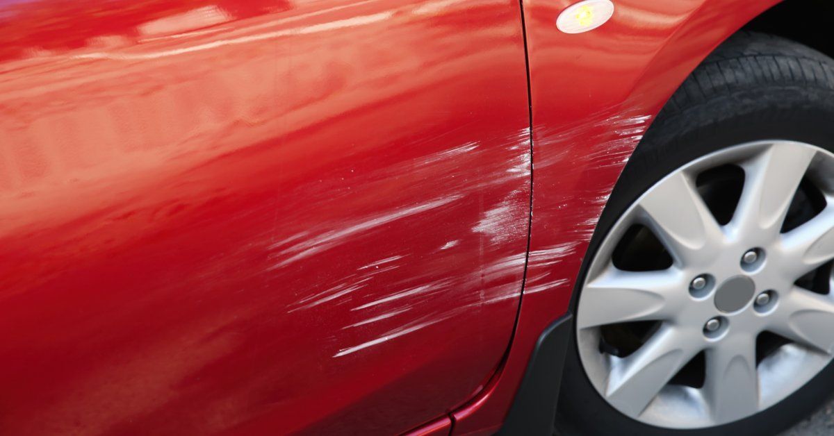 A red car with deep, white scratches near the passenger side door. The scratches extend toward the tire.