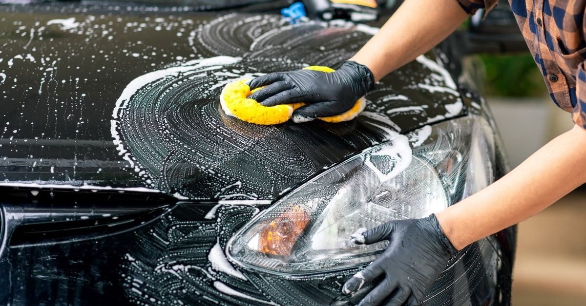 A person washing the front of their car with a yellow sponge. There is white soap across the hood of the vehicle.