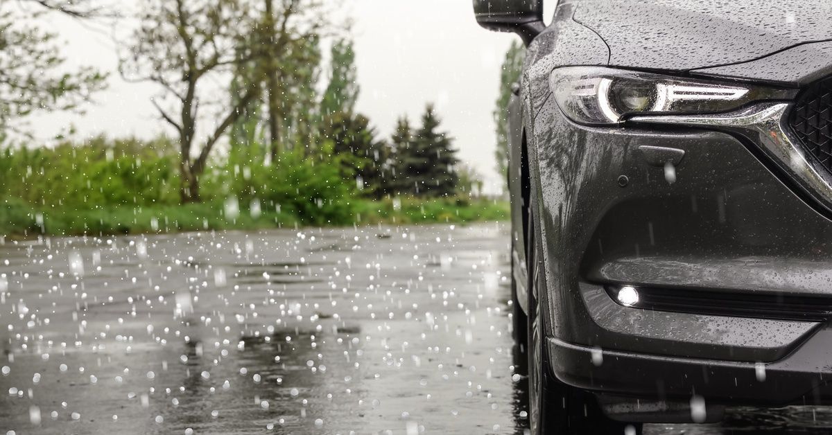 The front of a car with hail falling around it. The car is sitting in a parking lot with trees behind it.