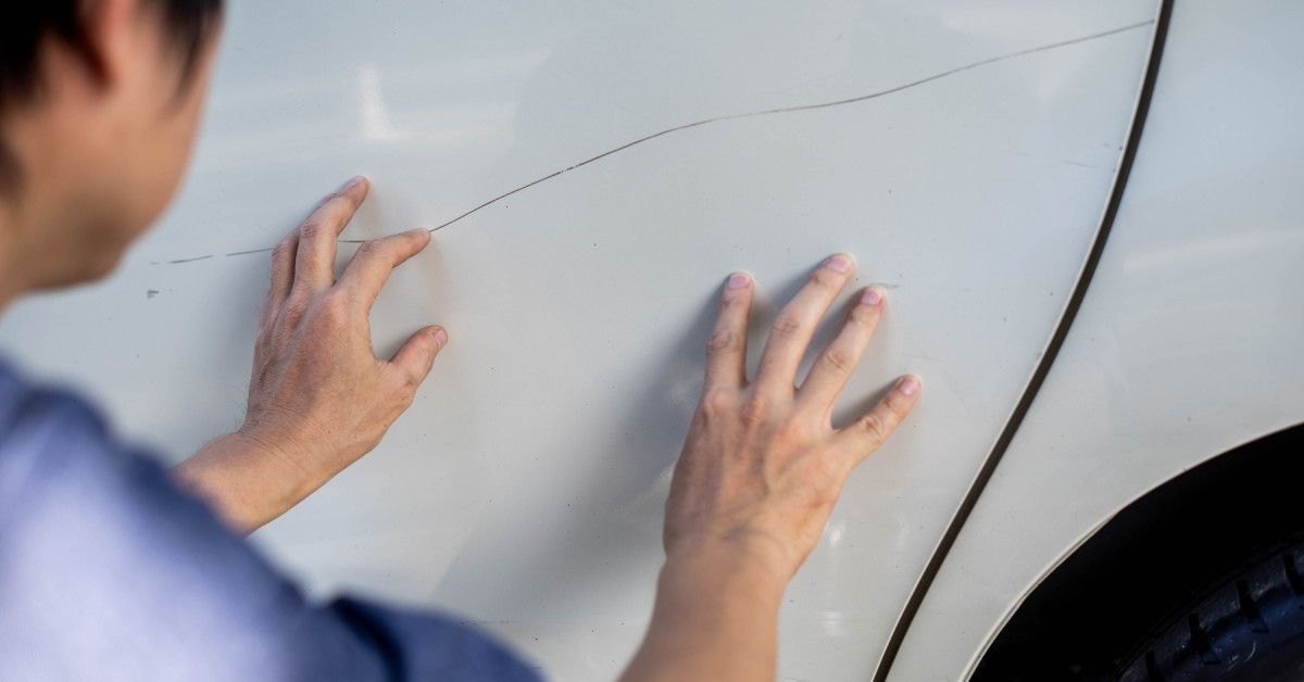 A man running both his hands along the door of a white sedan. There is an unsightly, long, horizontal scratch in the paint.