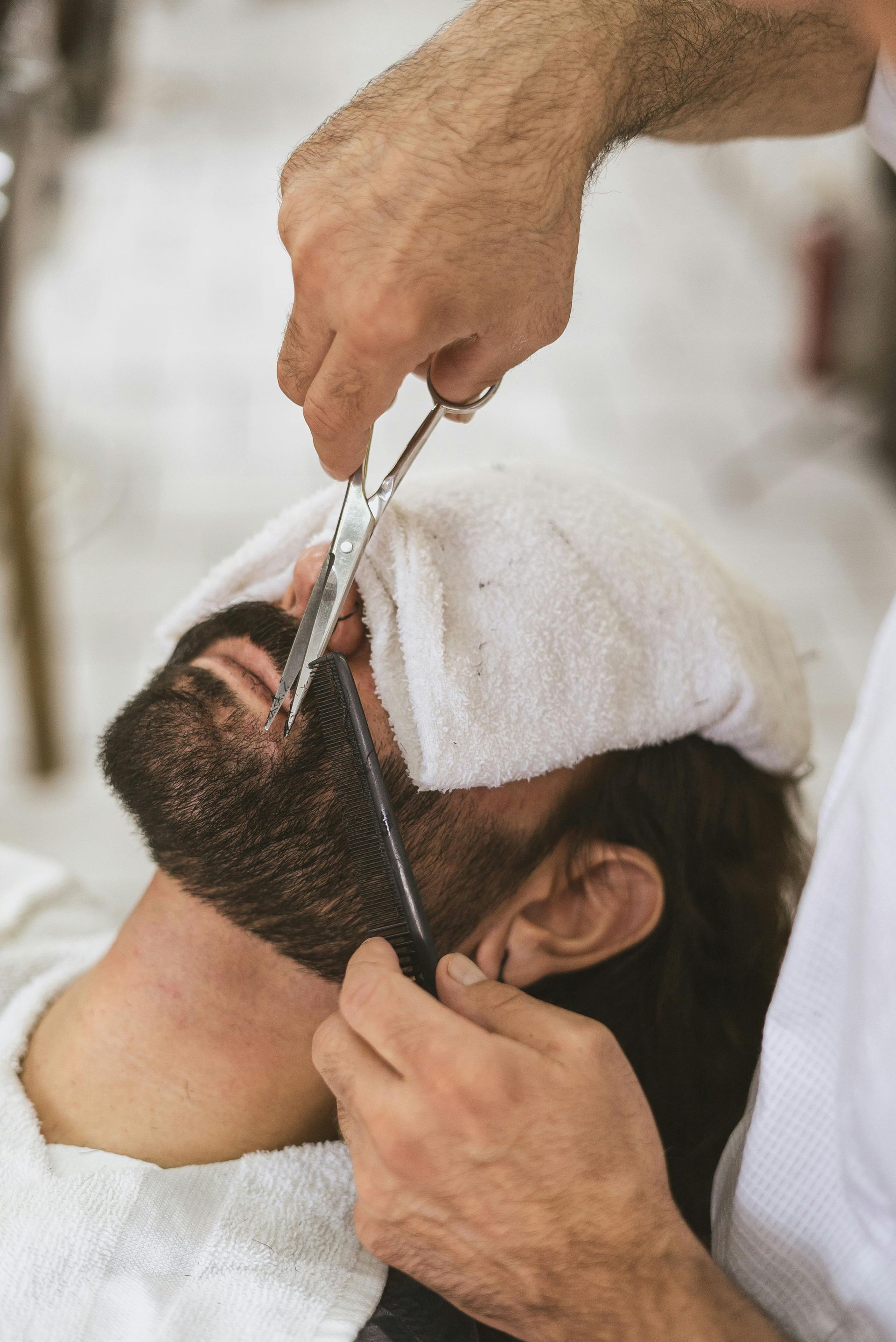 Barber trimming a man's beard with scissors and comb, using a towel around the face.