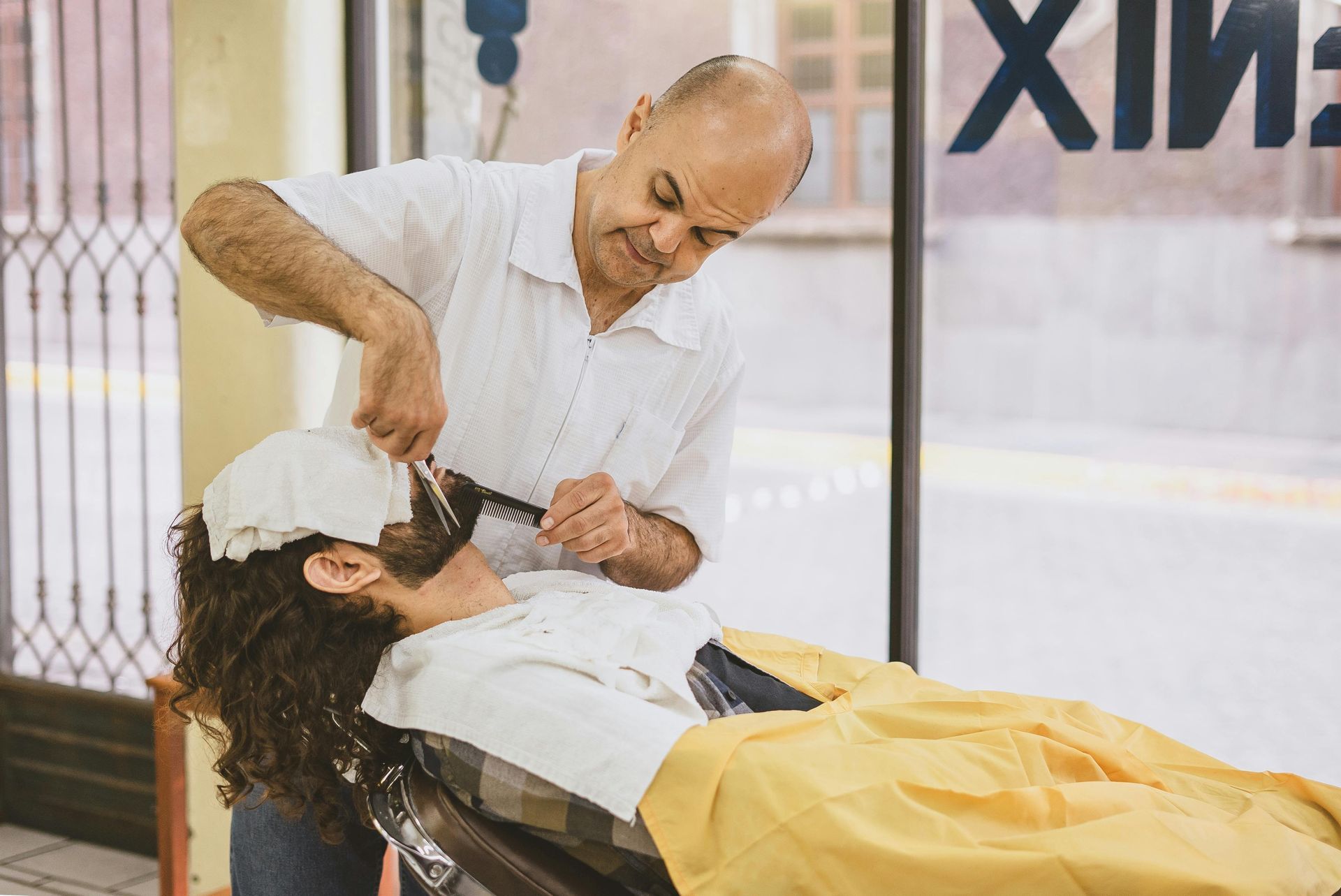 Barber shaves a client's face with a straight razor. The client is covered in a white cloth, lying down. Shop interior.