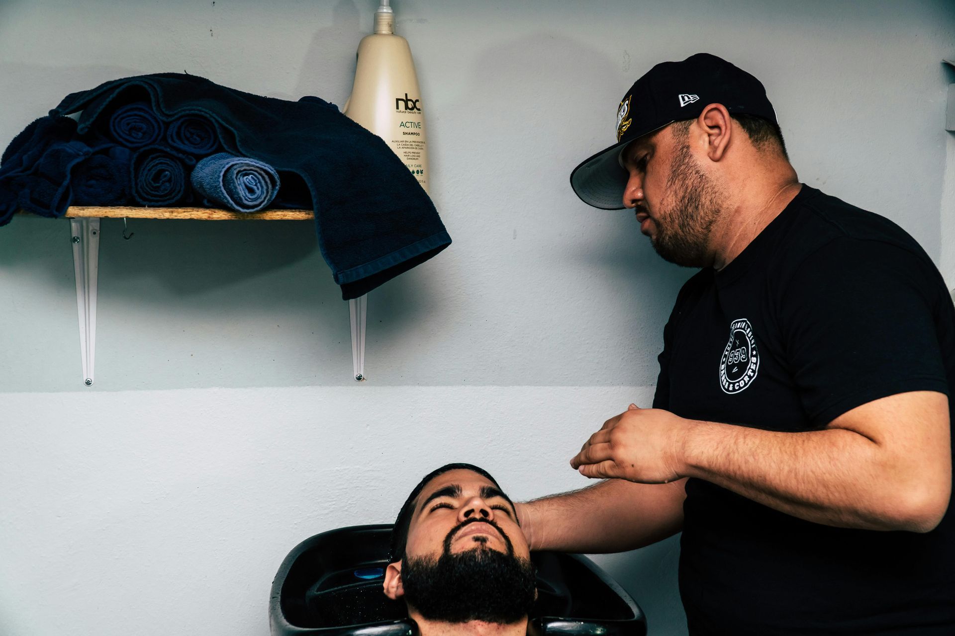 Barber washing client's hair at a salon. The barber is wearing a black shirt and cap.