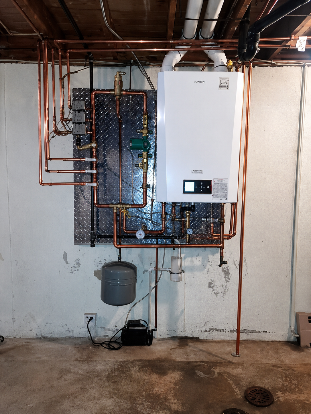 Wall-mounted boiler with copper pipes and a gray bucket in a basement utility room