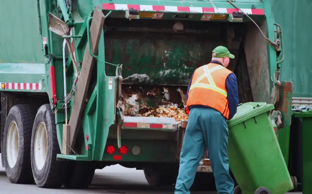 A man is loading a green trash can into a garbage truck.