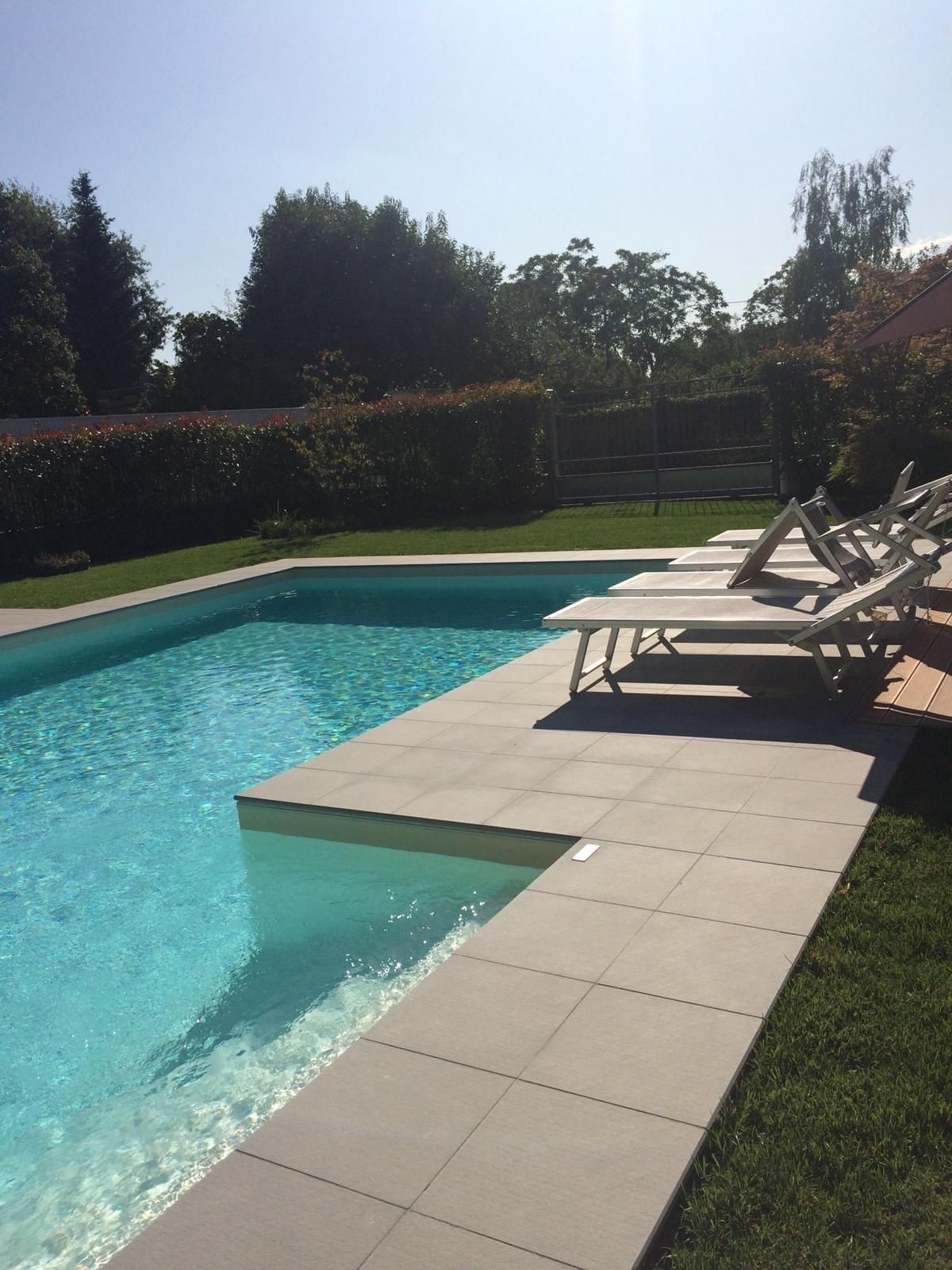 Piscina in giardino con acqua turchese, lettini prendisole e terrazza piastrellata accanto a un prato.