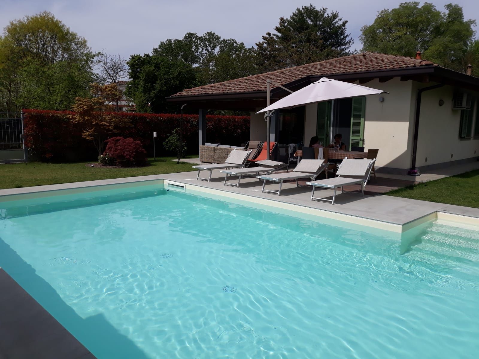 Piscina in giardino accanto a un patio con sedie a sdraio, ombrellone e una casetta