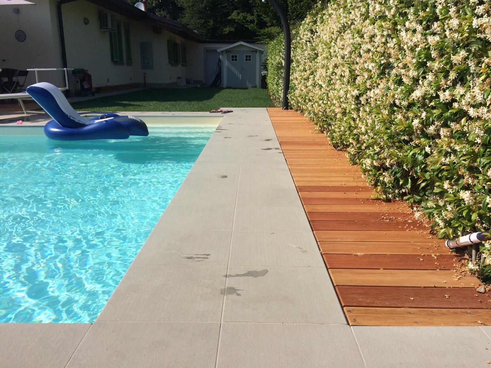 Piscina in giardino con vialetto in cemento e terrazza in legno accanto a una siepe verde.