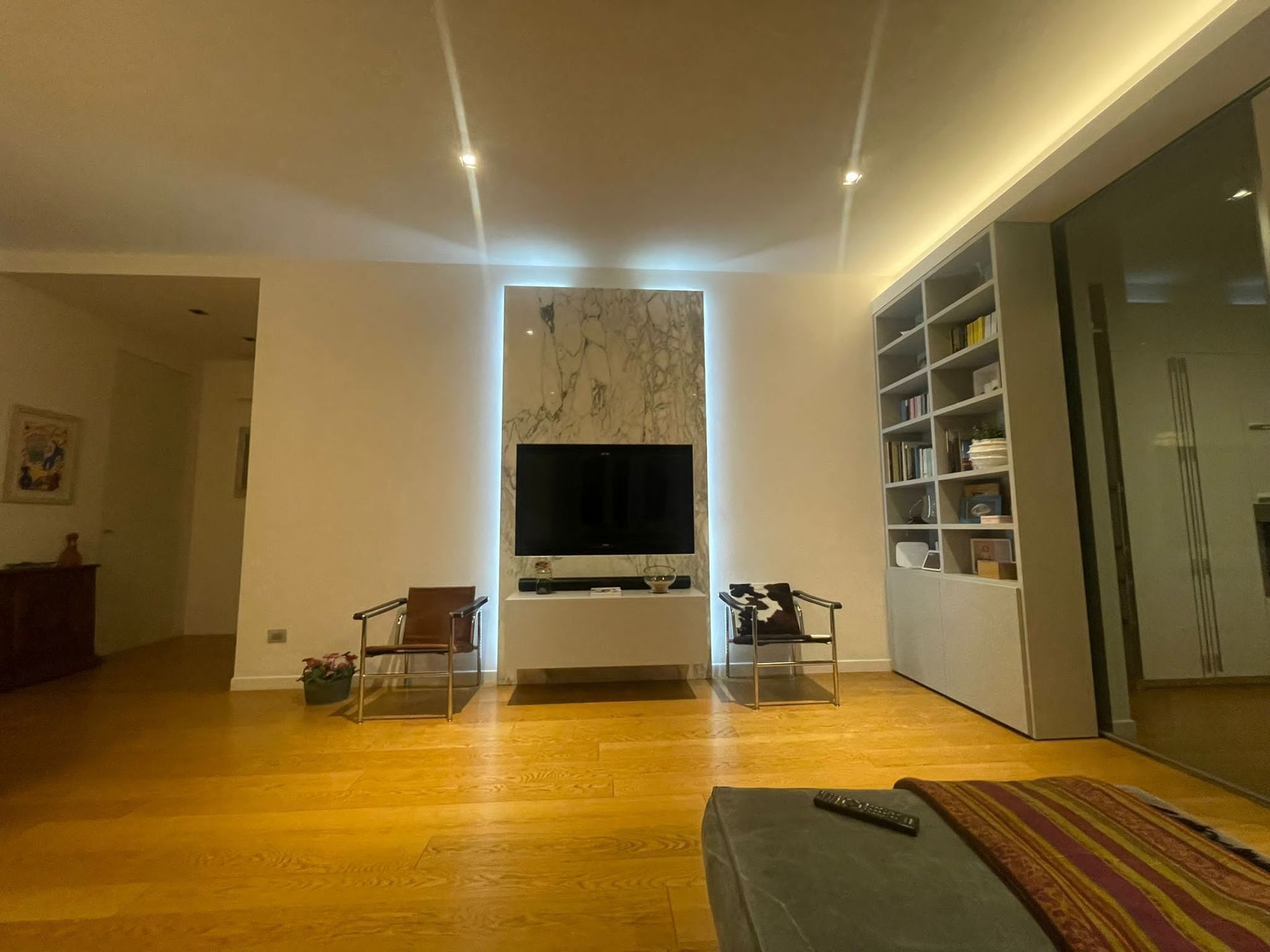 Soggiorno moderno con TV a parete, scaffali a muro, pavimento in legno e divani sotto luci a soffitto dai toni caldi.