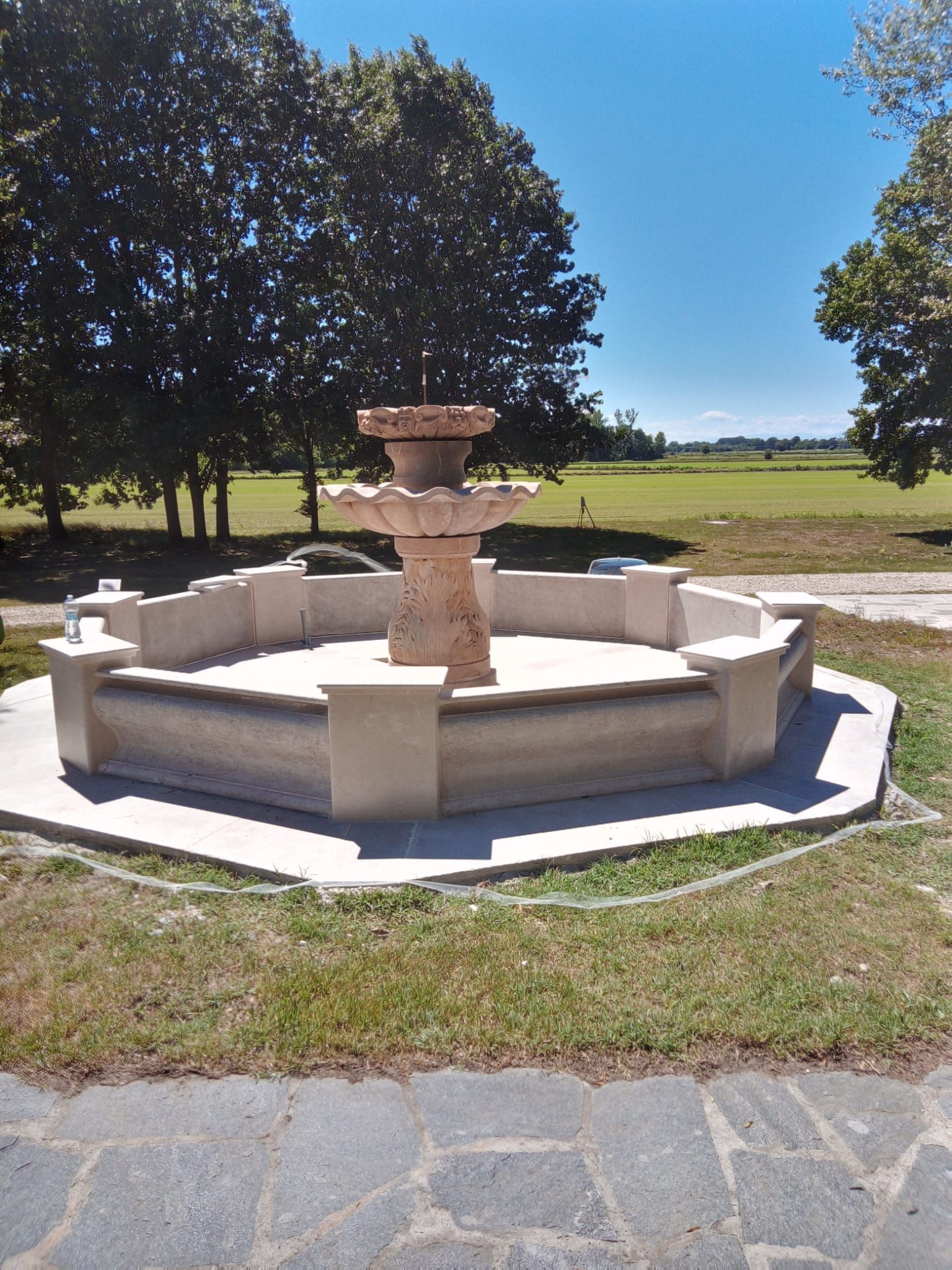 Fontana in pietra in un parco soleggiato e erboso, con alberi e campi aperti sullo sfondo.