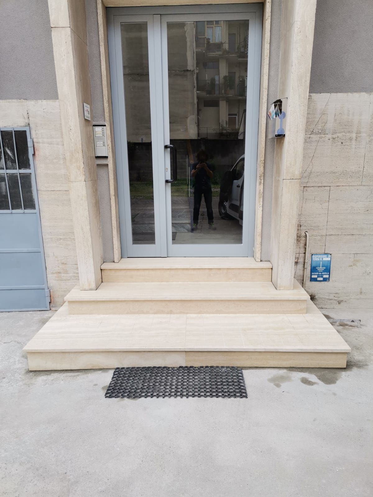 Ingresso dell'edificio con porta a vetri, gradini beige e zerbino nero su un vialetto di cemento.