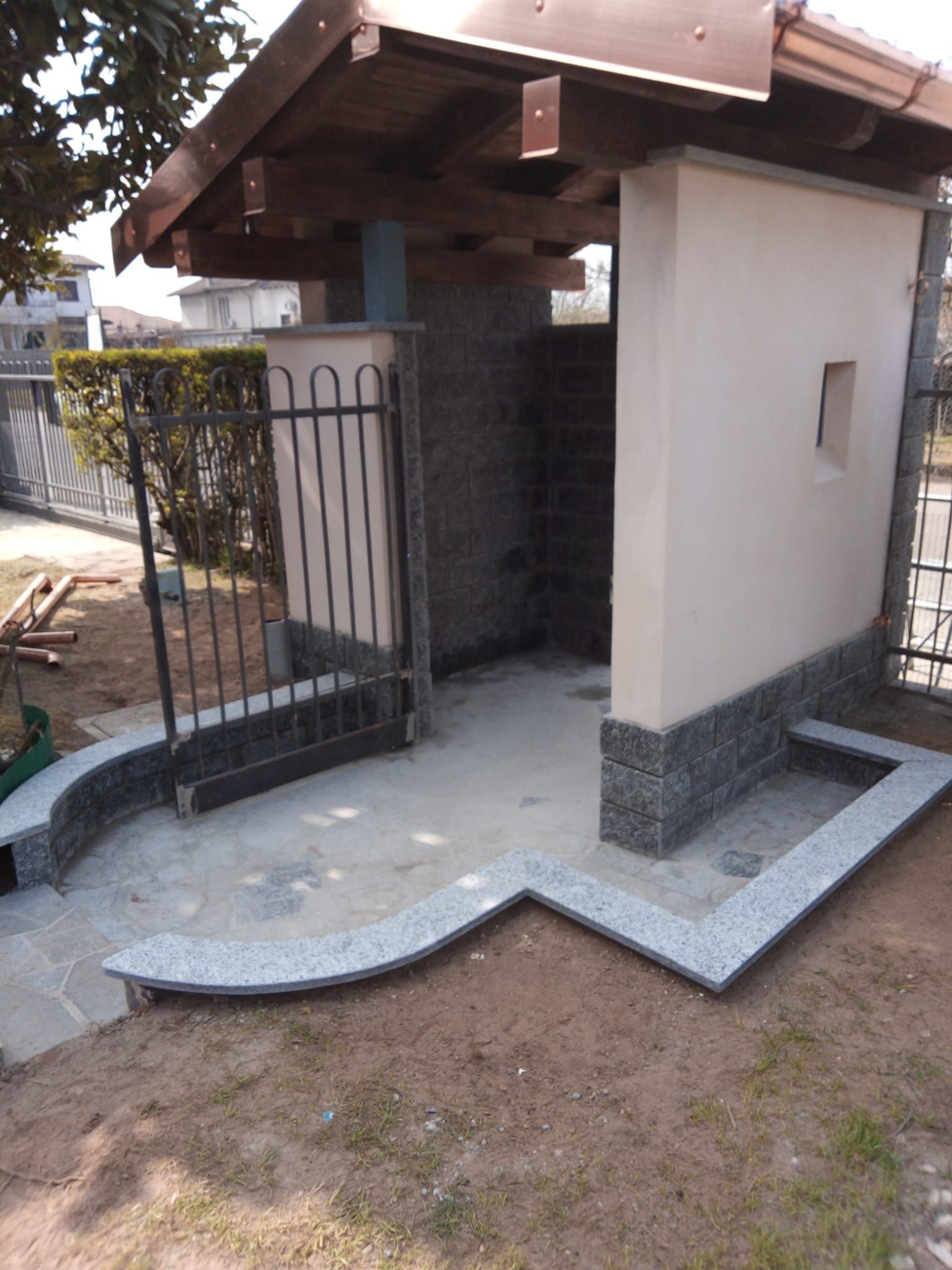 Piccolo ingresso di casa con portico coperto, cancello in metallo e rampa curva in pietra accanto a un cortile sterrato.