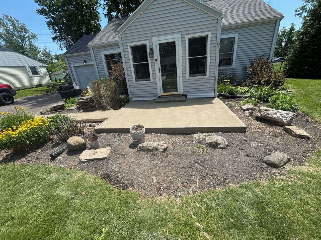 Front yard with a small house, concrete patio, and landscaped rock border with plants and stones.