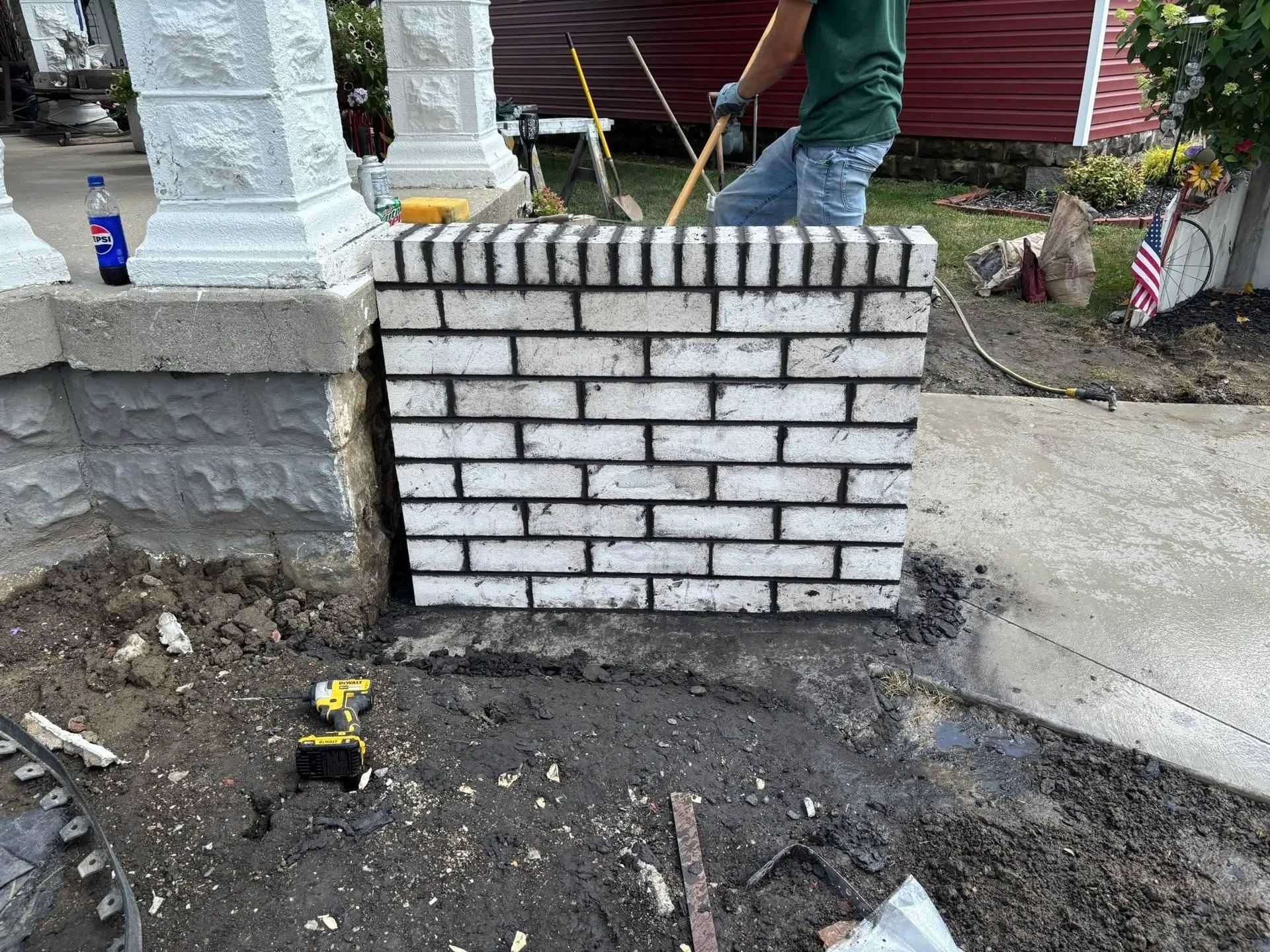Small brick retaining wall under construction beside a concrete walkway and dirt ground.