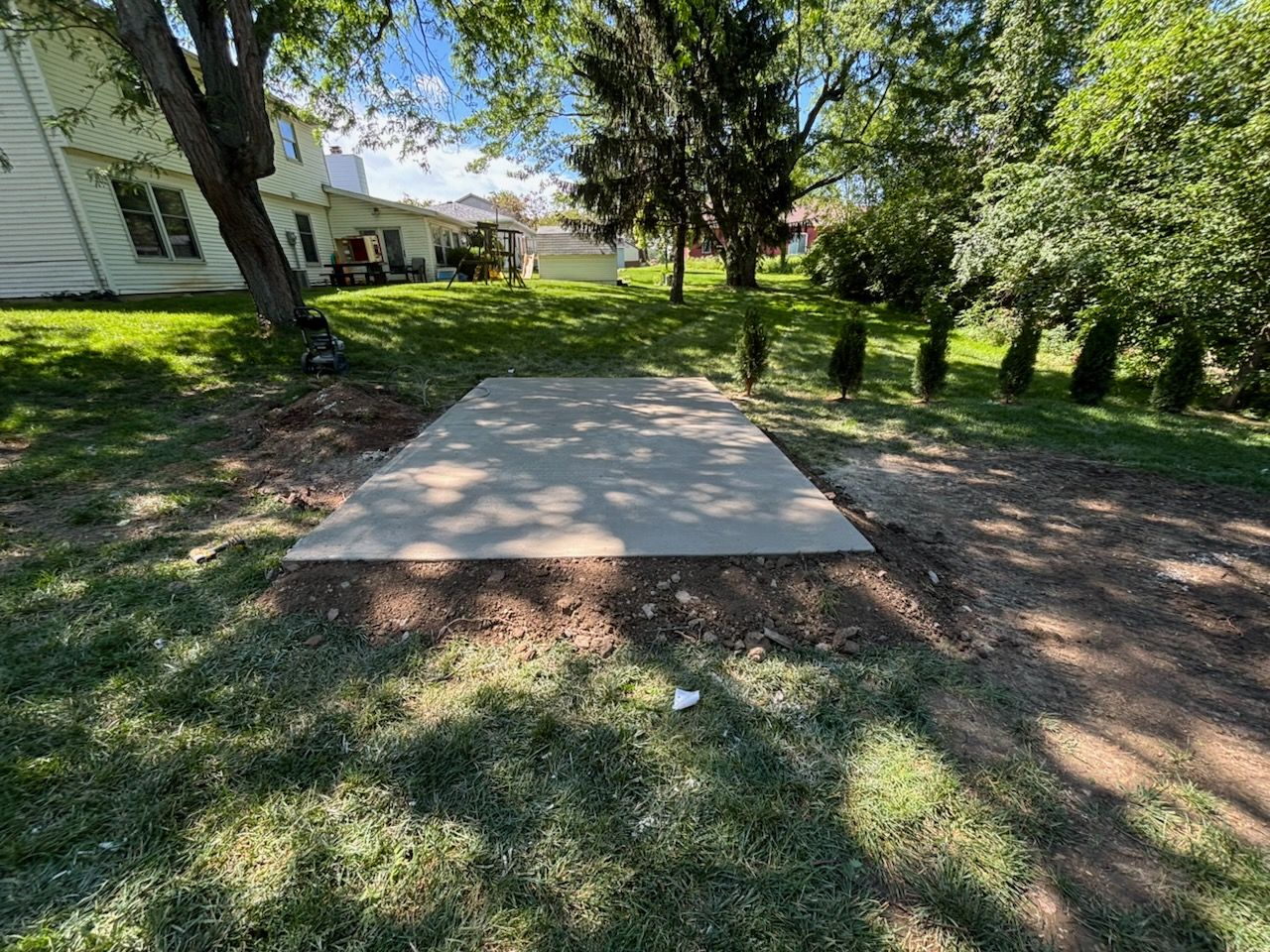 Concrete slab in a grassy, shaded yard beside a house, with trees and shrubs around it.
