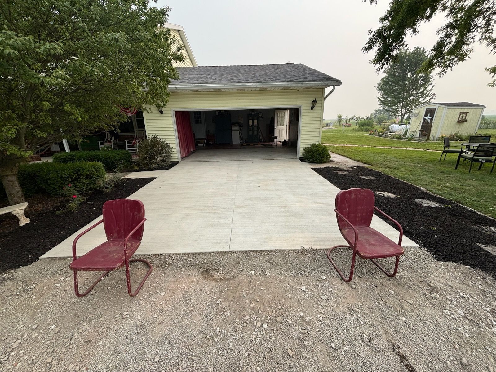 Driveway leading to a garage, with two red chairs on gravel in the foreground and trees on both sides.