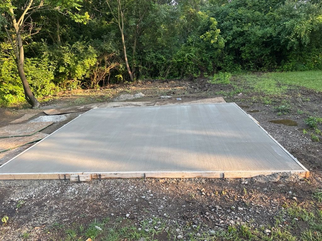 Concrete slab foundation in a wooded outdoor area, surrounded by gravel and dirt.