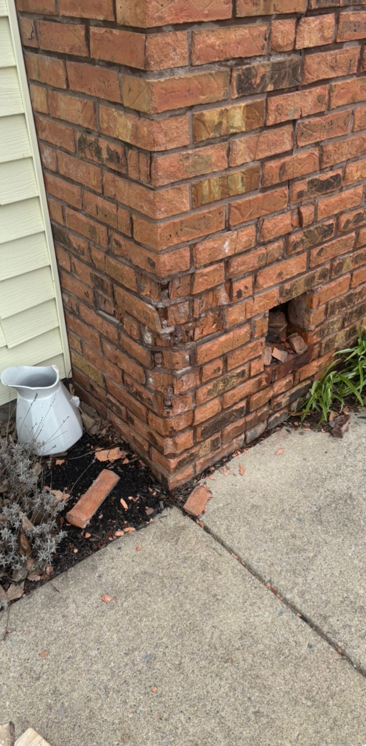 Damaged brick column by a sidewalk, with scattered bricks and a small white utility pipe beside it
