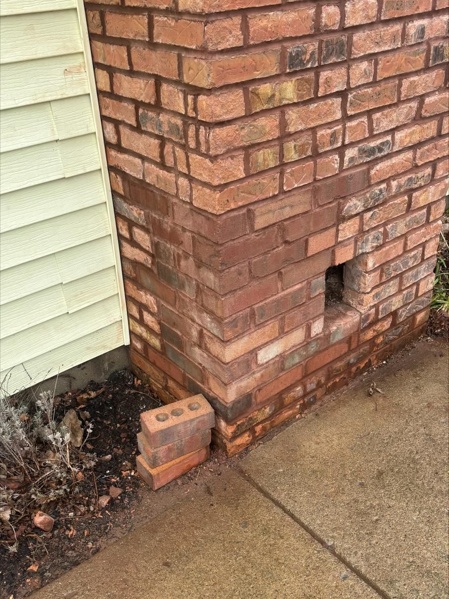Brick house corner with a small vent opening, adjacent siding, and a wet concrete walkway.