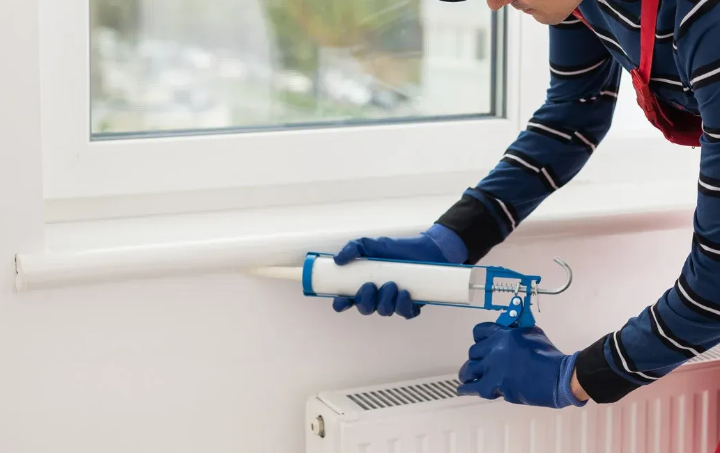 Person applying sealant to a white window sill with a caulking gun, wearing blue gloves