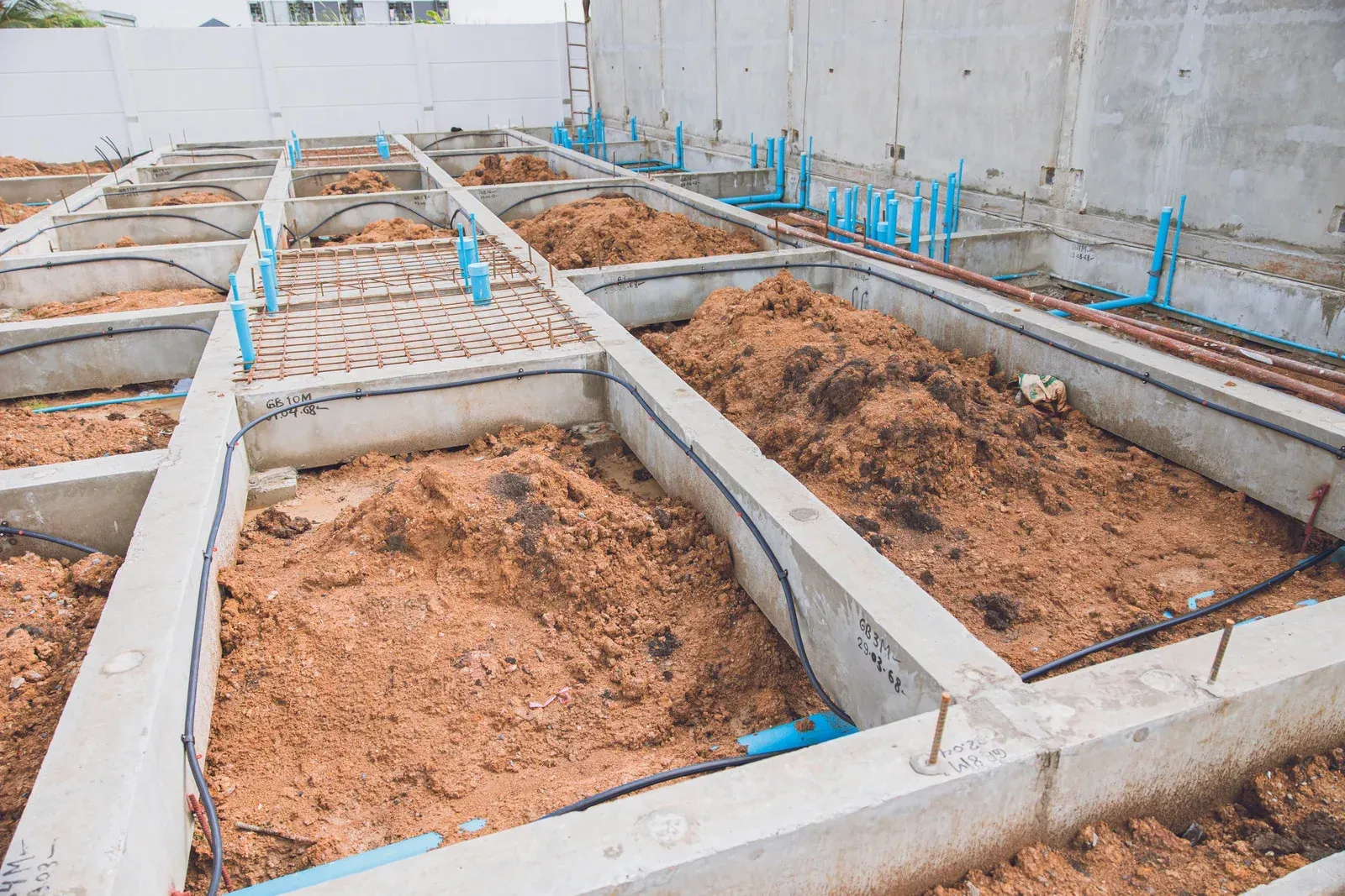 Concrete foundation footings with soil-filled trenches and blue plumbing pipes at a construction site