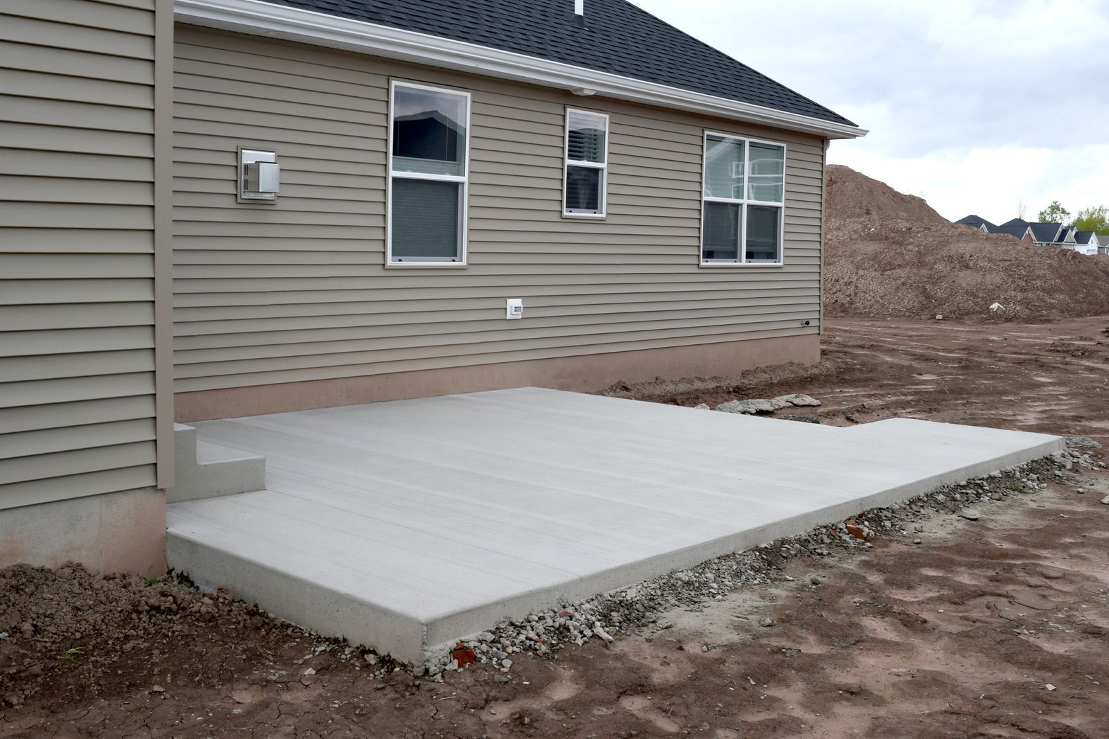 Concrete patio beside a beige house, with windows and dirt yard around it