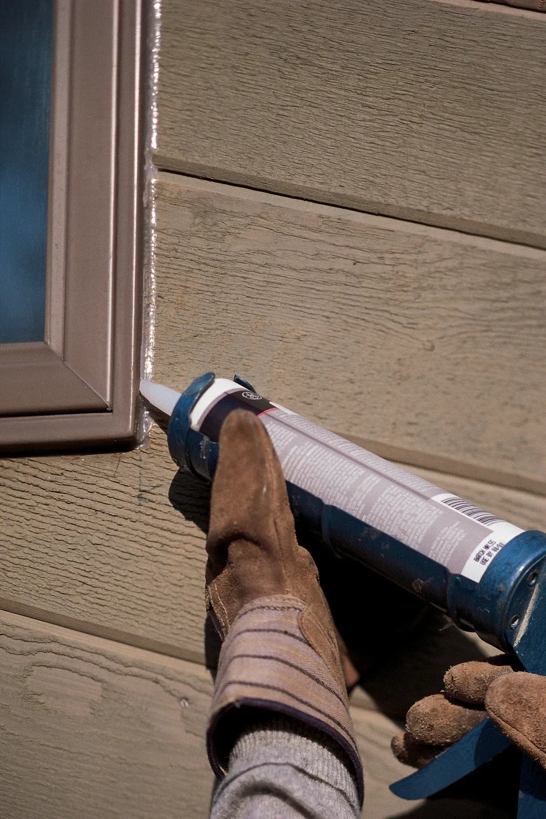 Gloved hand applying blue caulk to siding near a window frame
