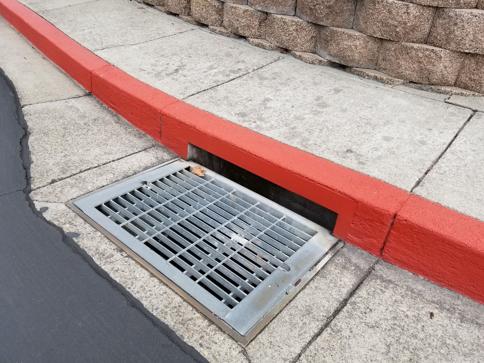 Street curb with a metal storm drain grate beside a red-painted curb and concrete sidewalk