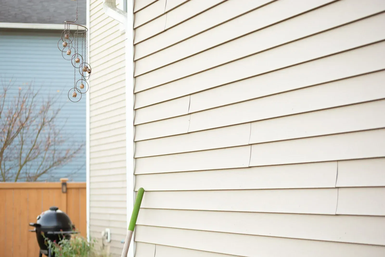 Side of a white house with beige siding, a broom leaning against it, and a grill near a wooden fence.