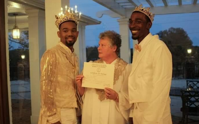 Two men in crowns flank a woman holding a certificate indoors at dusk.