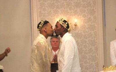 Two men in white ceremonial outfits face each other, wearing gold headpieces, in an ornate room.