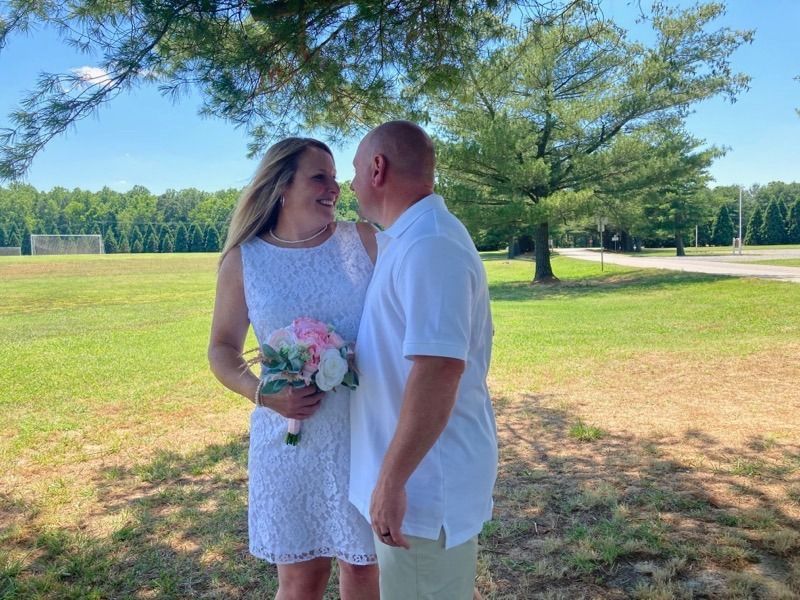 Couple standing in a sunny park, holding a bouquet and smiling at each other.