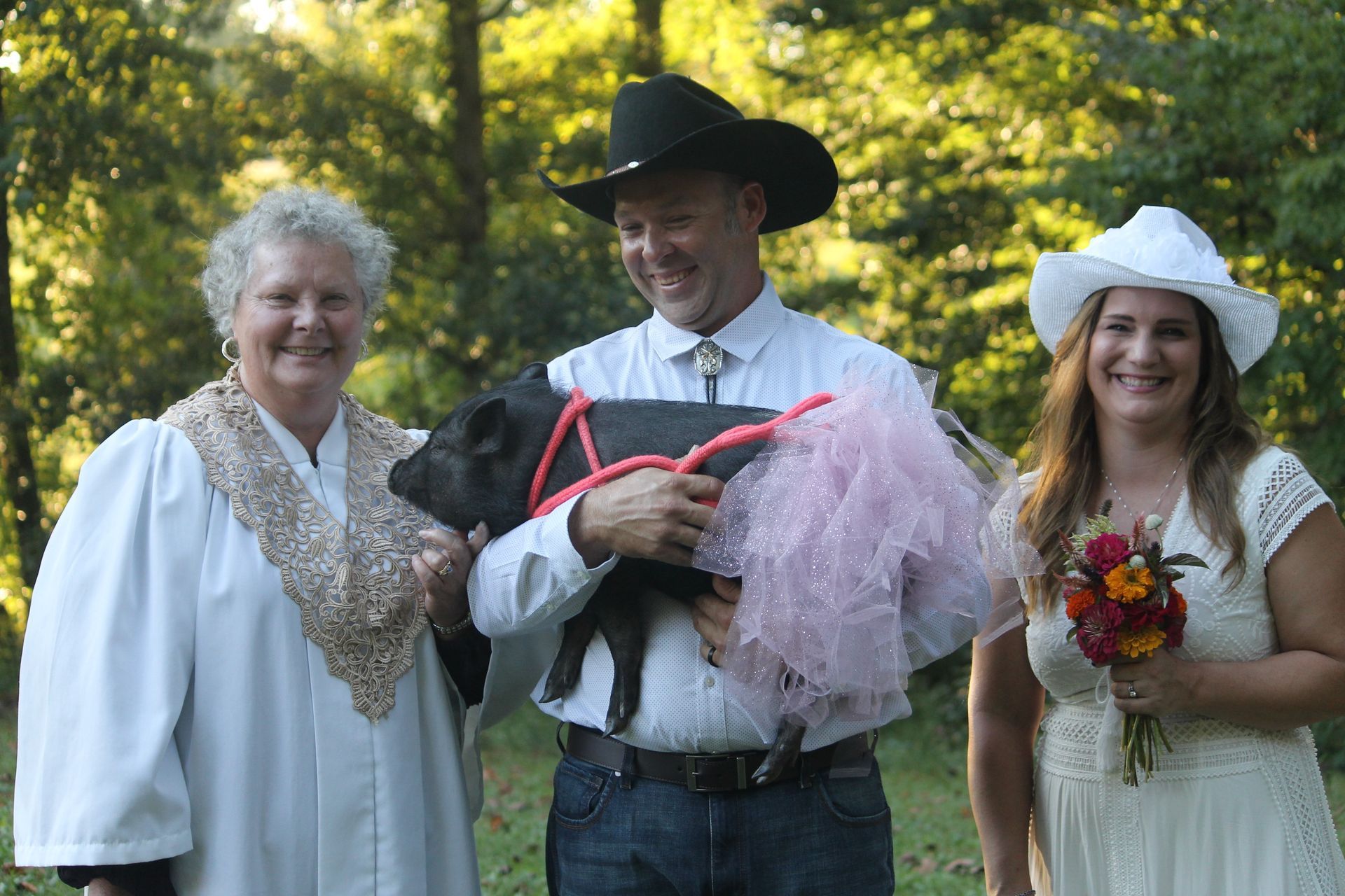 Three people in costumes outdoors, smiling and holding a pink tulle-wrapped item in a sunny wooded setting