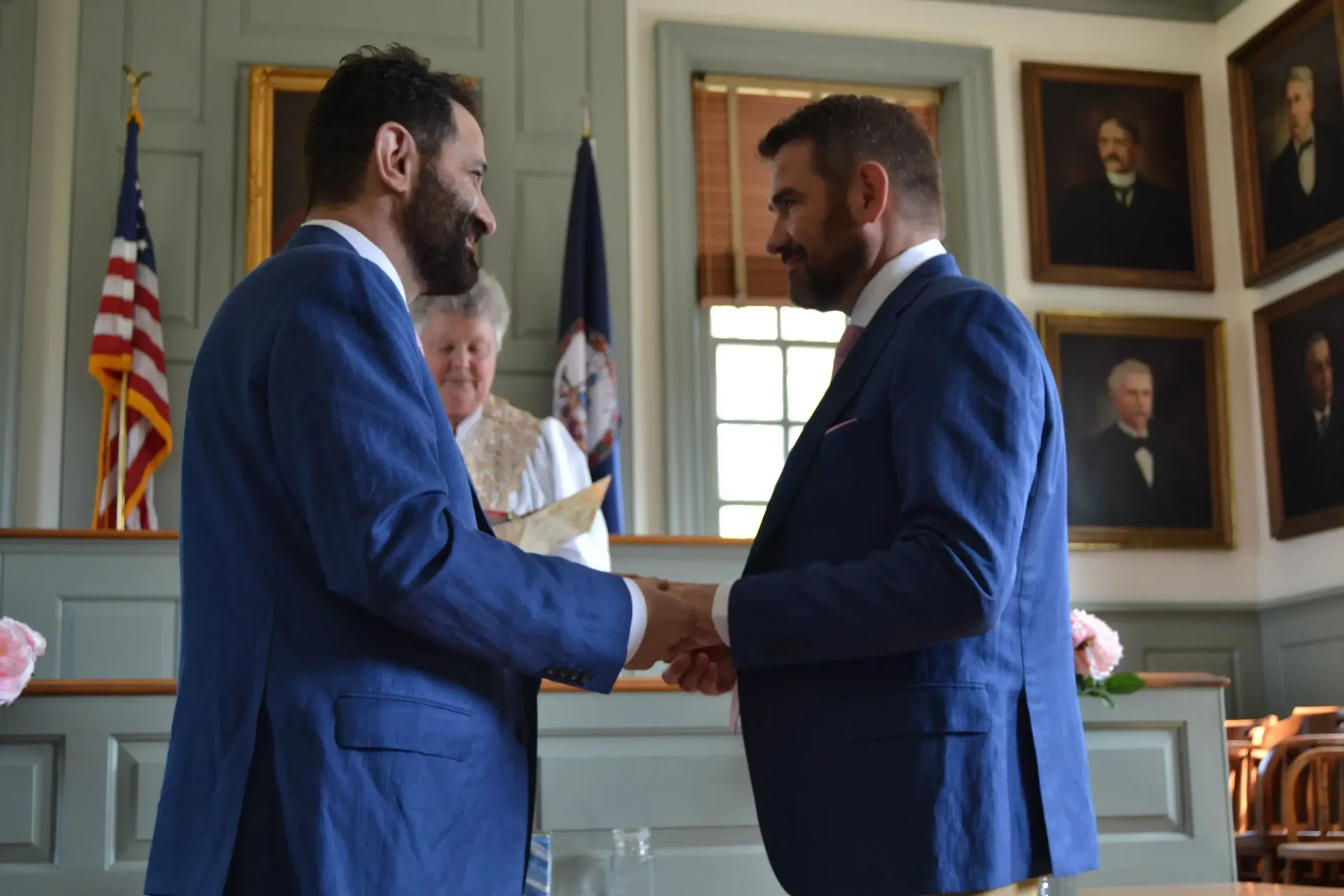 Two men in blue suits shake hands in a formal room with flags and framed portraits.
