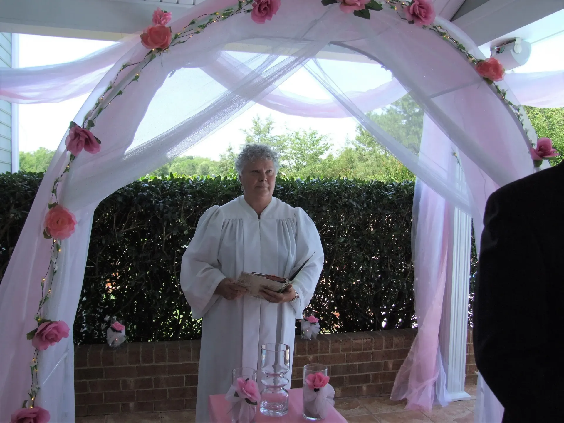 Person standing under a pink-draped floral arch at an outdoor ceremony, holding a book