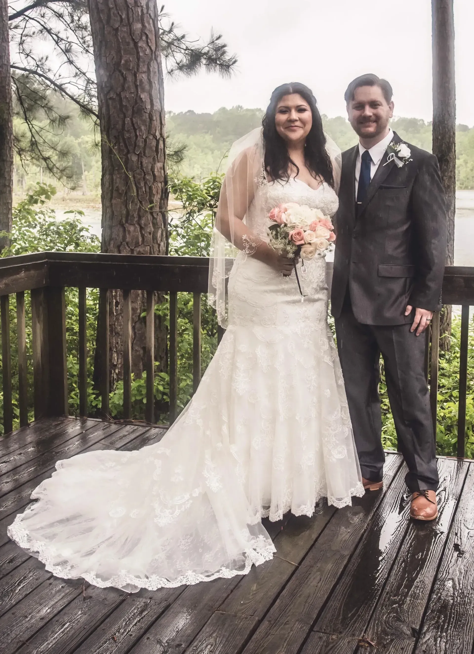 Bride and groom standing on a wooden deck, holding a bouquet in a rainy outdoor setting.
