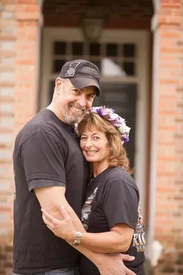 Smiling couple embracing outdoors in front of a brick building, the woman wearing a flower crown