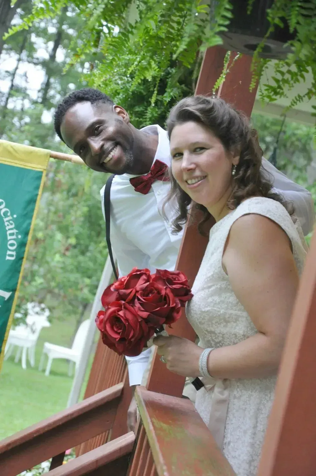 Smiling couple in wedding attire leaning on a porch railing, holding a red rose bouquet