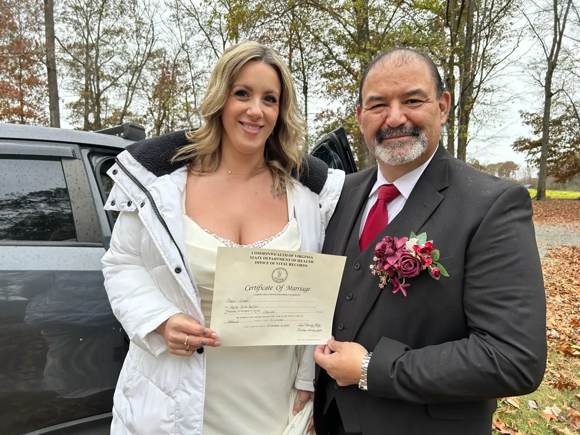 Couple posing outdoors, smiling and holding a certificate, with a black SUV and autumn trees behind them
