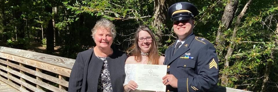 Three people smiling on a wooden bridge, holding a certificate in a sunny wooded park.