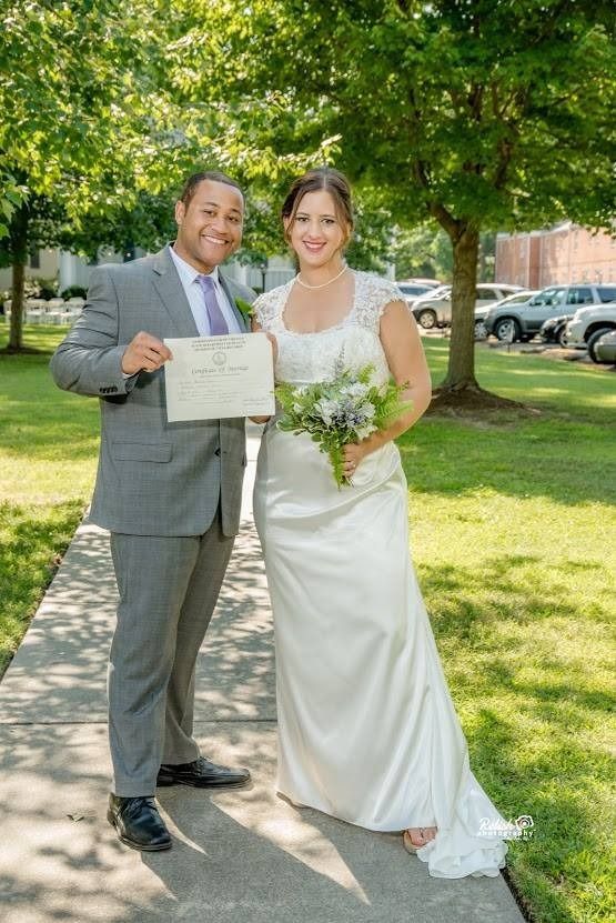 Two people in formal attire smiling outdoors, holding a marriage certificate and bouquet under a tree.