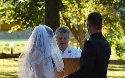 Outdoor wedding ceremony with bride in veil, officiant at podium, and groom facing her under trees