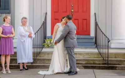 Couple in wedding attire embracing outside a red-doored church as guests watch nearby