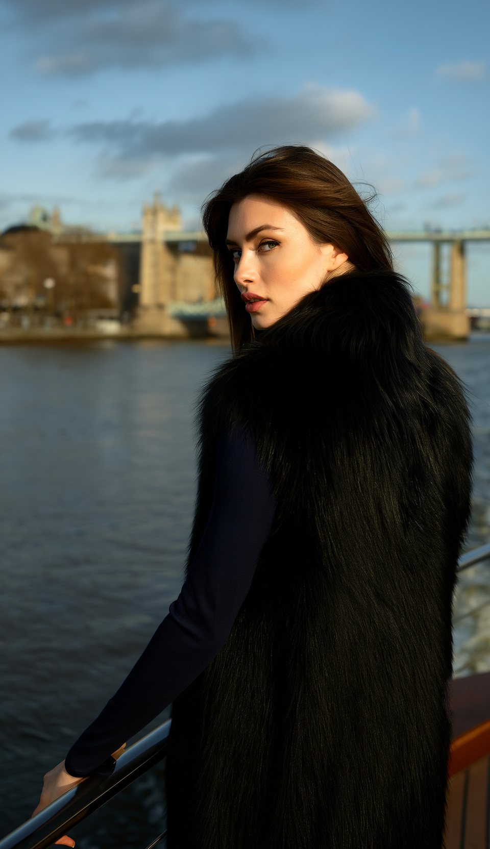 Donna con gilet e abito di pelliccia nera, in piedi su una barca con il Tower Bridge sullo sfondo.