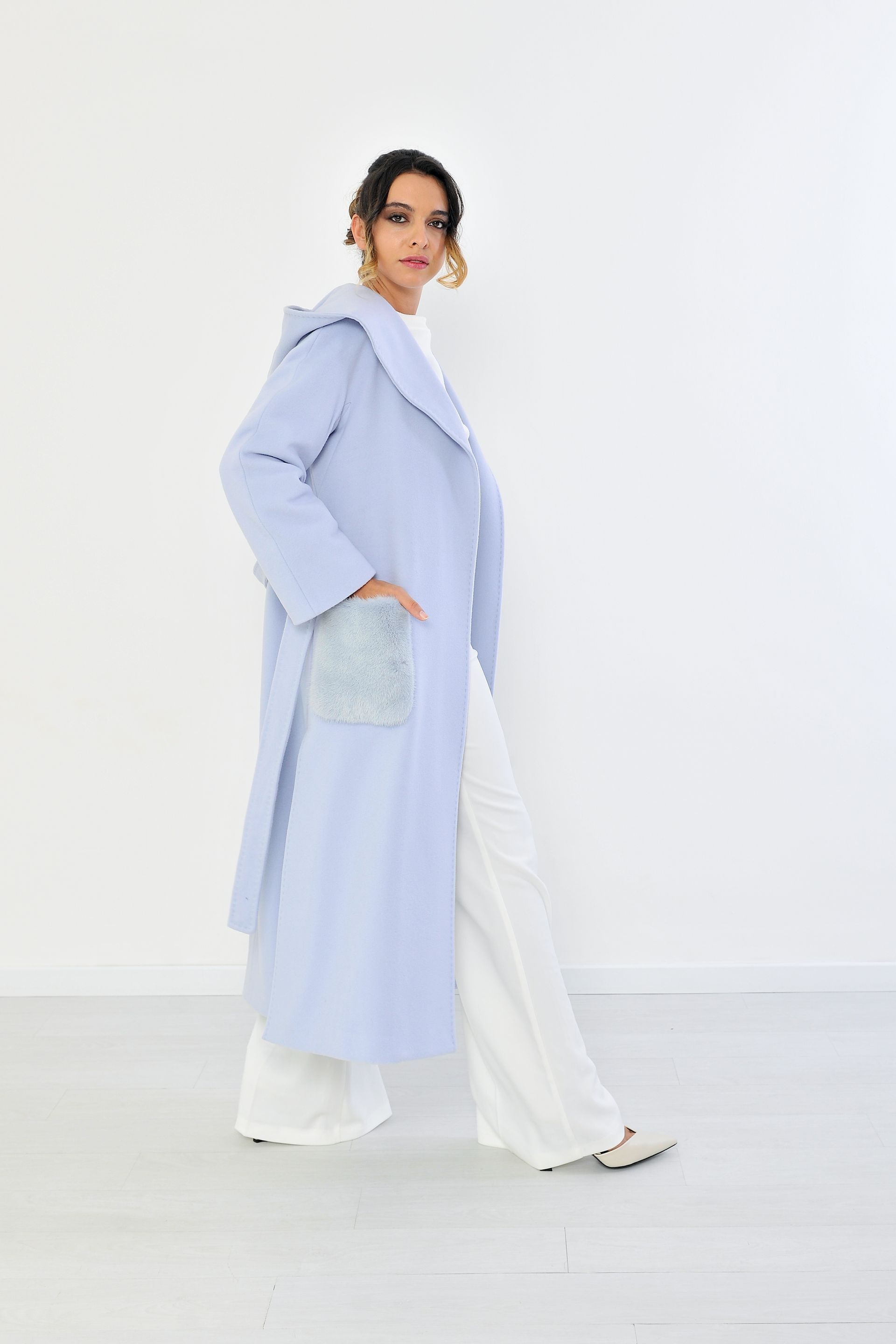 Donna con cappotto azzurro e pantaloni bianchi, in piedi con una mano in tasca contro un muro bianco.