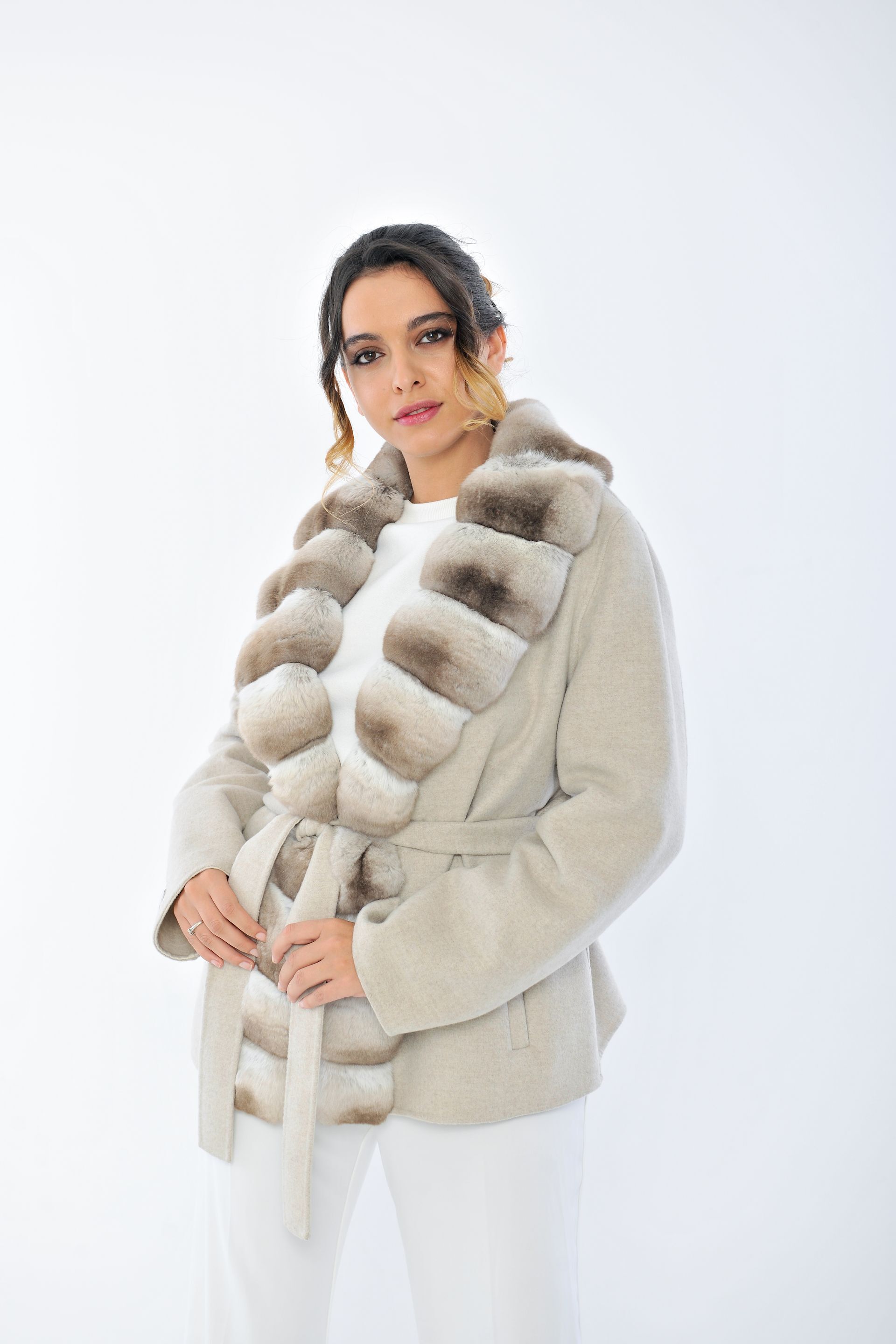 Una donna con pelliccia e pantaloni bianchi è in piedi su uno sfondo bianco, con le mani giunte.